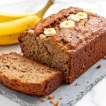 Freshly baked 2 banana bread recipe, with a golden brown crust, soft moist interior, and banana slices on top, served on a slate board with ripe bananas in the background.